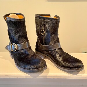 May include: A pair of black leather ankle boots with a fur-like texture. The boots have a buckle strap and a zipper on the side.