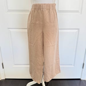 May include: Tan-colored wide-leg pants with an elastic waistband. The pants have a relaxed fit and a split detail on the sides. The fabric appears to be a soft, lightweight material, suitable for casual wear. The pants are displayed on a white mannequin.