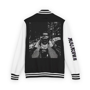 Puede incluir: Una chaqueta universitaria en blanco y negro con mangas blancas y puños y cuello de canalé a rayas negras. La espalda presenta un gran gráfico cuadrado de un motociclista con casco Shoei. El texto JUNGKOOK está impreso en la manga derecha.
