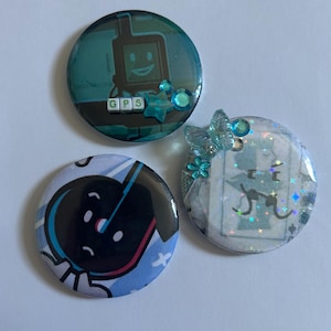 Op de afbeelding: Drie ronde buttons. Eén toont een talingkleurig personage met de tekst GPS en blauwe versieringen. Een andere toont een zwart-blauw personage met een droevige uitdrukking. De derde is een lichte button met vlinder- en steraccenten.