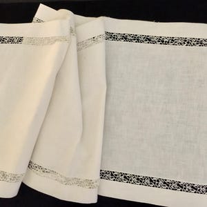 May include: A rectangular off-white table runner featuring a wide, solid fabric border surrounding a central panel. The runner is accented with an intricate, openwork lace trim that forms a decorative frame around the inner rectangular section.