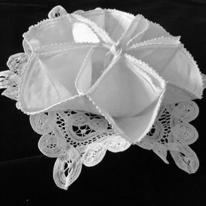 May include: A white fabric bowl with a star-shaped opening, constructed from folded panels with scalloped edges. The bowl rests upon a square white lace doily featuring intricate cutwork and floral patterns, creating a layered textile display.