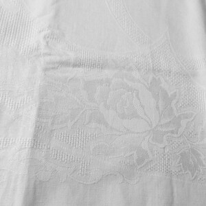 May include: A white textile fabric featuring a woven damask pattern. The design displays a prominent floral motif with detailed rose blossoms and leaves, integrated into a textured background with subtle geometric grid-like woven sections.