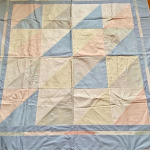 May include: A square quilt featuring a geometric pattern of triangles in soft pastel shades of light blue, pale pink, and cream. The design is composed of multiple square blocks arranged in a grid, surrounded by a solid light blue border frame.