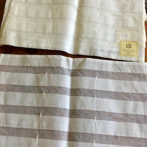 May include: A set of rectangular fabric placemats featuring a horizontal striped pattern in shades of white and light gray. Each placemat includes a small square label in the bottom corner with the text Wilton Court and a crown logo icon.