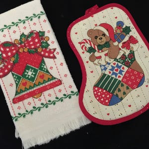 May include: A white rectangular hand towel with a fringed edge featuring a red and green bell graphic, alongside a cream-colored, rounded pot holder with a red border displaying a teddy bear wearing a Santa hat and holding a candy cane.