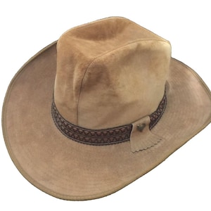 May include: A tan suede cowboy hat featuring a wide brim and a pinched crown. The hat is accented with a dark brown decorative band that has a geometric pattern and a small leather tab with a heart-shaped metal embellishment on the side.