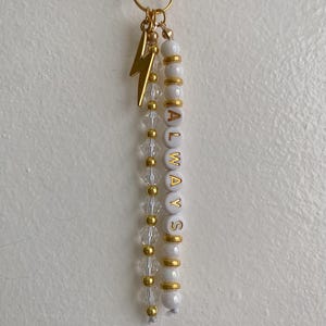 May include: A gold-toned keychain featuring two dangling strands attached to a ring. One strand consists of alternating clear faceted beads and small gold beads with a gold lightning bolt charm. The other strand features white beads with the gold text ALWAYS.