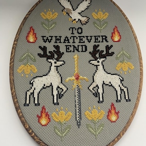 May include: An oval cross-stitch embroidery in a wooden hoop featuring a gray fabric background. The design includes two white deer, a central sword, a flying bird, yellow flowers, and flames, with the black text TO WHATEVER END.