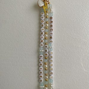 May include: A beaded keychain featuring three strands of white, light blue, and gold-toned beads. The white beads display the text LIGHTNING STRIKES, DANCING THROUGH, and THE. The keychain includes a gold-toned lightning bolt charm and a white heart charm.