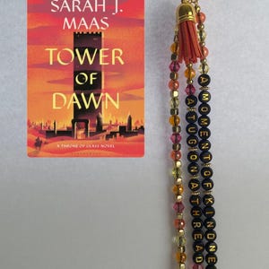 May include: A keychain featuring a gold-tone lobster clasp, a small orange tassel, and three beaded strands. One strand displays black letter beads spelling A MOMENT OF KINDNESS A TUG ON A THREAD, alongside a book cover for Tower of Dawn by Sarah J. Maas.