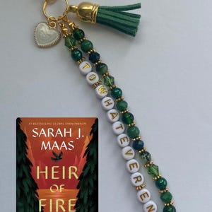 May include: A gold-toned keychain featuring a green tassel, a white heart charm, and two strands of green beads. One strand includes white letter beads spelling TO WHATEVER END. Beside it is the book cover for Heir of Fire by Sarah J. Maas.