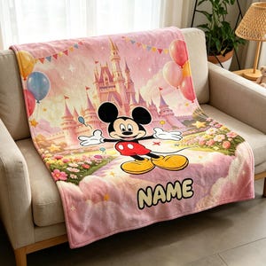Könnte beinhalten: Eine weiche, rosafarbene Fleecedecke mit einer farbenfrohen Illustration von Mickey Mouse vor einem großen Schloss. Das Design zeigt schwebende Luftballons, ein Blumenbeet und das Wort NAME in großen, fetten Buchstaben mit dunkler Umrandung.