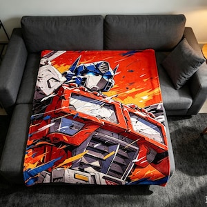 Puede incluir: Una manta rectangular de forro polar con un gráfico vibrante y estilizado del personaje Optimus Prime. El diseño muestra la armadura metálica roja y azul del robot, una rejilla de camión detallada y ojos azules brillantes sobre un fondo naranja.