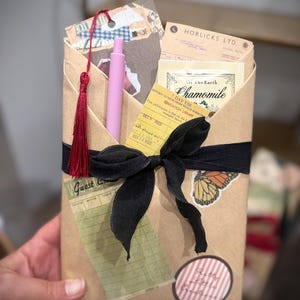 May include: A brown paper-wrapped package tied with a black ribbon, featuring a pink pen, a red tassel, a Horlicks Ltd. document, a chamomile tea bag, a yellow library card, a green guest check, a butterfly sticker, and a round striped sticker.
