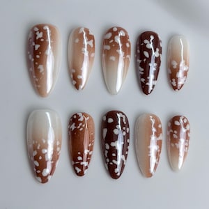 May include: A set of ten almond-shaped press-on nails featuring a glossy finish. Each nail displays a gradient transition from a deep chocolate brown to a lighter tan or cream base, accented with irregular white speckled patterns across the surface.