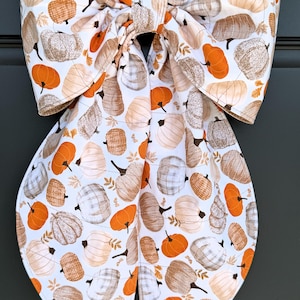 May include: A large fabric bow featuring a repeating pattern of pumpkins in shades of orange, white, and beige on a white background. The bow has two wide, long tails and a central knot, constructed from a printed textile with visible stitching.