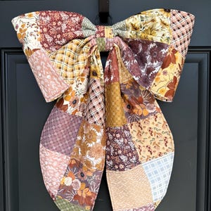 May include: A large decorative bow constructed from a patchwork of various fabric patterns in shades of mustard yellow, burnt orange, brown, cream, and muted green. The fabric features floral prints, plaid designs, and small geometric motifs.
