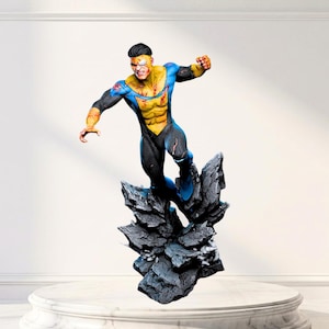 May include: A detailed action figure of a superhero in a blue and yellow suit with black accents, depicted in a dynamic flying pose above a jagged, dark gray rocky base. The figure features visible battle damage, including red blood markings.