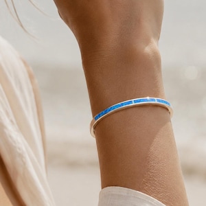 Może przedstawiać: Srebrna bransoletka typu bangle z ciągłą inkrustacją z jasnoniebieskich, prostokątnych kamieni. Metalowa obręcz ma polerowane wykończenie i smukły profil, otaczając nadgarstek serią żywych niebieskich segmentów osadzonych w srebrnej ramie.