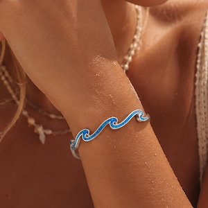 May include: A silver cuff bracelet featuring a continuous wave design inlaid with bright blue crushed opal. The metal band follows a fluid, undulating pattern that wraps around the wrist, showcasing a polished finish and vibrant blue texture.