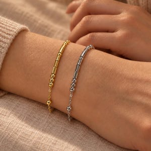 May include: Two delicate bracelets, one in gold tone and one in silver tone, featuring a sequence of small round beads and cylindrical tube beads arranged along a thin metal chain. The bracelets are worn side by side on the wrist.