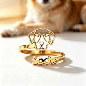 Peut inclure: Une bague ouverte dorée présentant le contour stylisé d'une tête de chien à une extrémité et le prénom Sadie en écriture cursive à l'autre extrémité. L'anneau en métal est poli et réfléchissant, avec un design en ligne continue pour le visage.