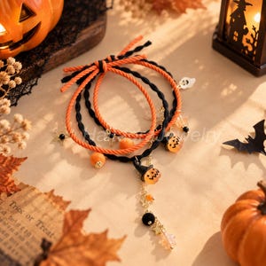 Op de afbeelding: Een set van drie gelaagde armbanden met oranje en zwarte gedraaide koorden. De armbanden zijn versierd met kleine bedels, waaronder een wit spookje, oranje vilten pompons, goudkleurige sterren en een bungelende bedel van een pompoen met hoed.