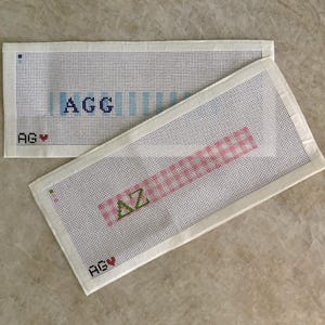 May include: Two rectangular white needlepoint canvases with grid patterns. The top canvas features the blue text AGG and a small red heart. The bottom canvas displays the green text AZ, a pink grid pattern, and a small red heart. Both have AGV printed.
