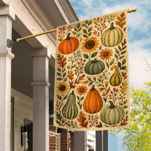 May include: A rectangular garden flag featuring a cream-colored background with a repeating pattern of orange and green pumpkins, gourds, sunflowers, and autumn leaves. The flag is mounted on a gold-toned metal pole attached to a white porch pillar.