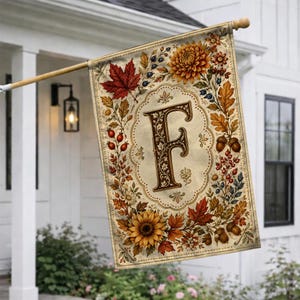 May include: A rectangular garden flag featuring a large, ornate letter F in the center, surrounded by a decorative border of autumn-themed elements including orange sunflowers, red maple leaves, brown acorns, and small red berries on a cream background.