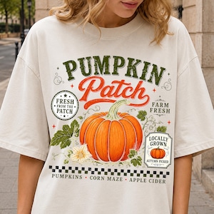 May include: An off-white t-shirt featuring a large orange pumpkin graphic with green leaves and vines. The text reads PUMPKIN PATCH, FRESH FROM THE PATCH, FARM FRESH, LOCALLY GROWN AUTUMN PICKED, PUMPKINS, CORN MAZE, and APPLE CIDER in various fonts.