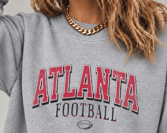 May include: A heather gray crewneck sweatshirt featuring the red text ATLANTA in a collegiate font, with the word FOOTBALL and a small football graphic printed in black underneath the main lettering.