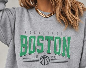 May include: A heather gray crewneck sweatshirt featuring the green text "BOSTON" in a collegiate font, with "BASKETBALL" above and "EST 1946" below, accompanied by a black basketball graphic and horizontal lines on the chest.