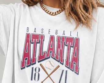 May include: A white short-sleeved t-shirt featuring a graphic print with the text BASEBALL ATLANTA 1871. The design includes red block lettering for ATLANTA, blue text for BASEBALL and 1871, and an illustration of a baseball and two crossed bats.