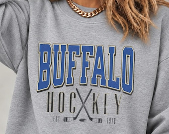 May include: A heather gray crewneck sweatshirt featuring the blue text BUFFALO in a collegiate font above the black text HOCKEY with crossed hockey sticks and a puck graphic, including the text EST 1970 at the bottom of the design.