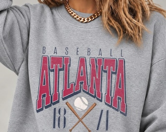 May include: A heather gray crewneck sweatshirt featuring a graphic print with the text BASEBALL ATLANTA 1871. The design includes large red block lettering, a white baseball, and two crossed brown wooden bats centered on the chest.