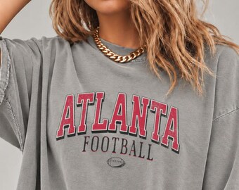 May include: A charcoal gray short-sleeve t-shirt featuring the word ATLANTA in red block letters with a black outline, the word FOOTBALL in smaller black letters, and a small graphic of a football centered below the text.