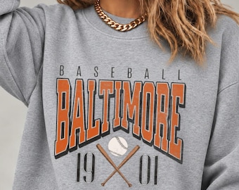 May include: A heather gray crewneck sweatshirt featuring the text BASEBALL BALTIMORE 1901 in orange and black lettering. The graphic includes a white baseball and two crossed brown wooden bats centered below the city name.
