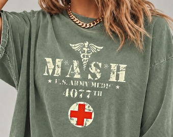 May include: An olive green short-sleeve t-shirt featuring a distressed white graphic with the text MASH, U.S. ARMY MEDIC, 4077TH, a caduceus symbol, and a circular red cross emblem centered on the chest area of the garment.
