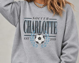 May include: A heather gray crewneck sweatshirt featuring a graphic print with the text SOCCER CHARLOTTE EST 2019. The design includes a black and white soccer ball centered above two blue laurel branches, with the word CHARLOTTE in large dark blue letters.