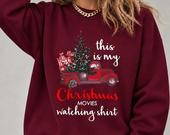 May include: A maroon long-sleeve sweatshirt featuring a graphic of a red pickup truck carrying a Christmas tree and gifts. The text reads: this is my Christmas MOVIES watching shirt. The design includes white and red lettering.