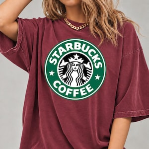 May include: A maroon short-sleeved t-shirt featuring a large, circular green and white Starbucks Coffee logo printed on the chest. The garment has a crew neckline and a relaxed, loose-fitting silhouette with dropped shoulder seams.
