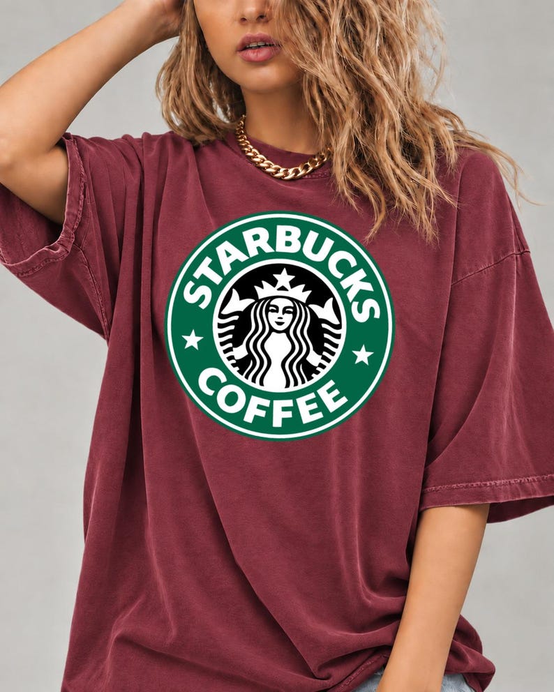 May include: A maroon short-sleeved t-shirt featuring a large, circular green and white Starbucks Coffee logo printed on the chest. The garment has a crew neckline and a relaxed, loose-fitting silhouette with dropped shoulder seams.
