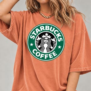 May include: An oversized, burnt orange t-shirt featuring a large, circular green and white Starbucks Coffee logo printed on the center of the chest. The shirt has short sleeves and a crew neckline, with a visible seam along the shoulder and sleeve.