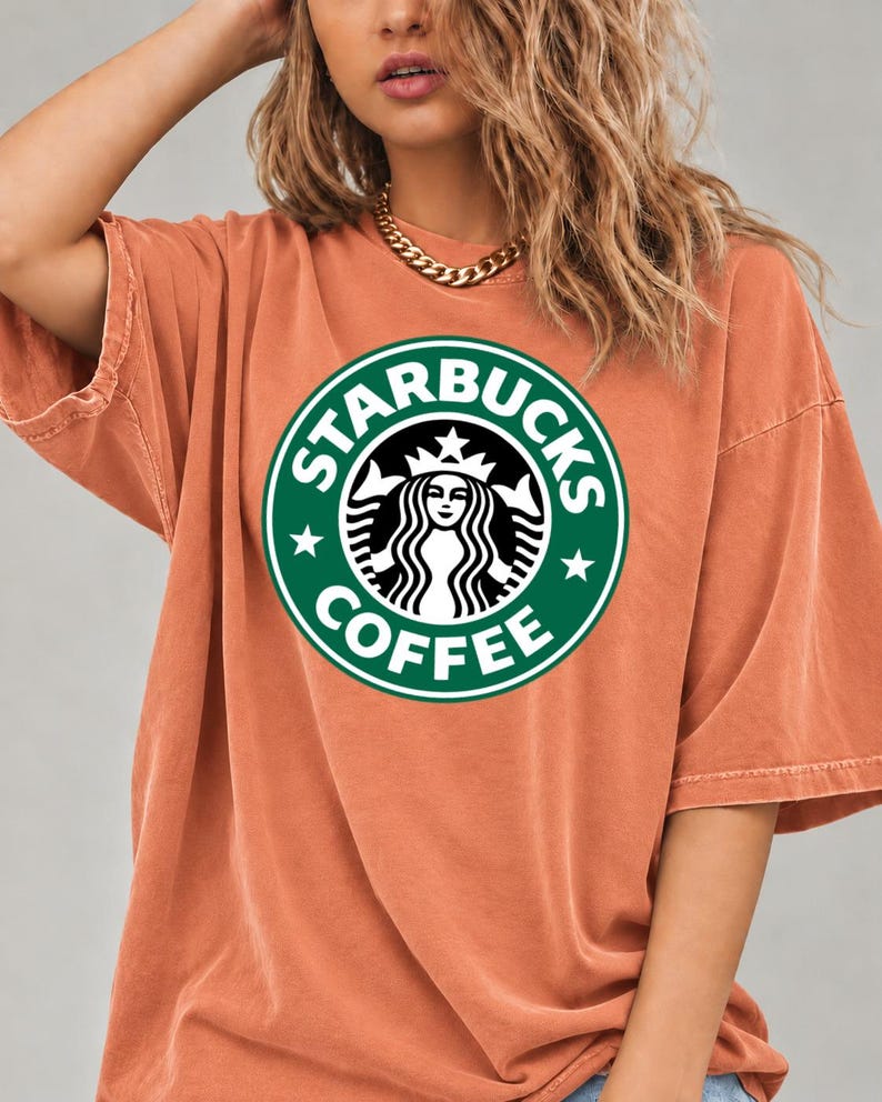 May include: An oversized, burnt orange t-shirt featuring a large, circular green and white Starbucks Coffee logo printed on the center of the chest. The shirt has short sleeves and a crew neckline, with a visible seam along the shoulder and sleeve.