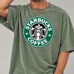 May include: An olive green short-sleeve t-shirt featuring the circular Starbucks Coffee logo in white and green on the chest. The shirt has a crew neckline and a relaxed fit, paired with a gold-toned chain necklace worn around the neck.