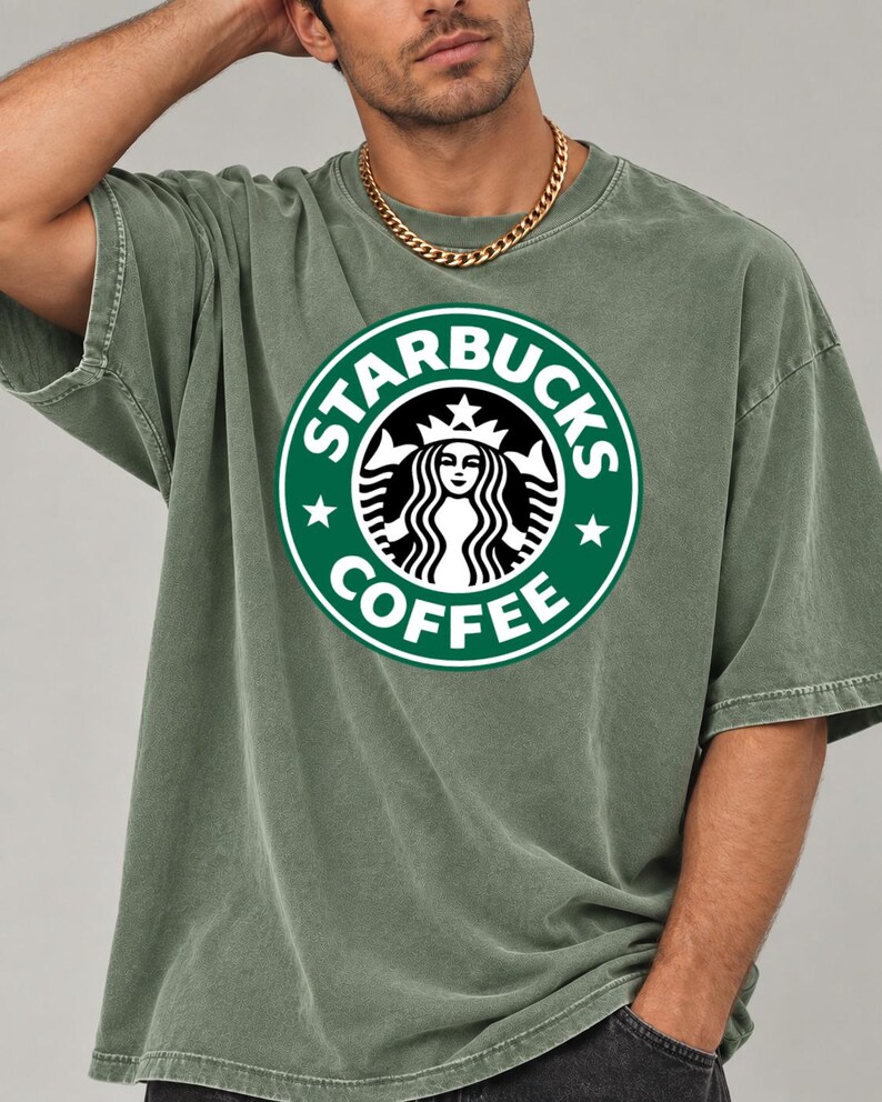 May include: An olive green short-sleeve t-shirt featuring the circular Starbucks Coffee logo in white and green on the chest. The shirt has a crew neckline and a relaxed fit, paired with a gold-toned chain necklace worn around the neck.