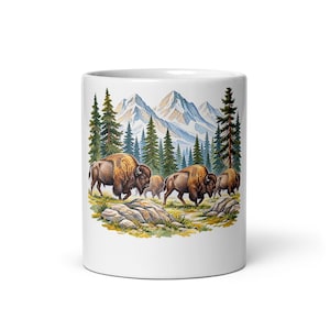 May include: A white ceramic mug featuring a colorful illustration of a herd of brown bison grazing in a grassy field. The background displays a dense forest of green pine trees set against a backdrop of tall, snow-capped mountain peaks.
