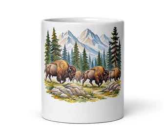 May include: A white ceramic mug featuring a colorful illustration of a herd of brown bison grazing in a grassy field. The background displays a dense forest of green pine trees set against a backdrop of tall, snow-capped mountain peaks.